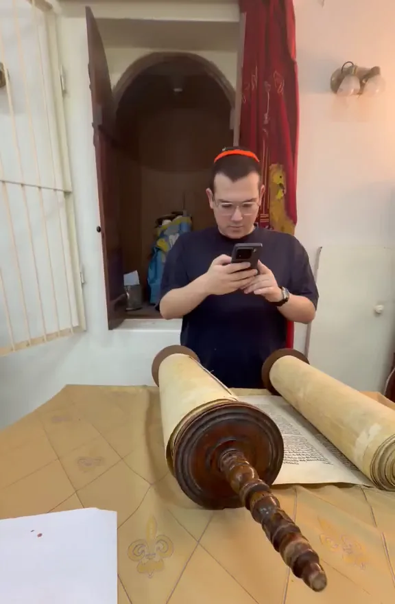 Mati with his phone in front of the open sefer on the bimah